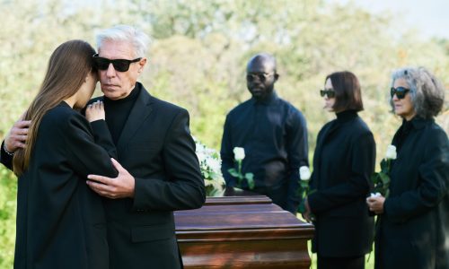 Grieving mature man in black suit embracing his mourning daughter while expressing his sympathy for loss of dear friend or family member